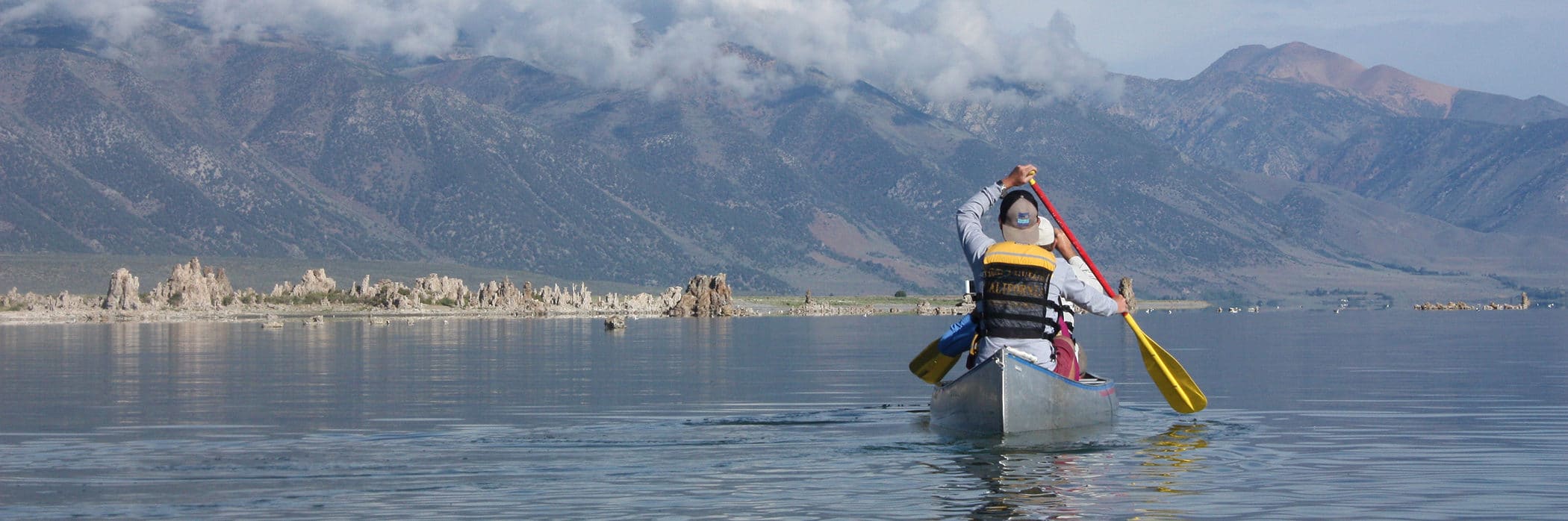 View from behind a person paddling a canoe on Mono Lake with tufa towers and tall mountains shrouded in clouds in the distance.