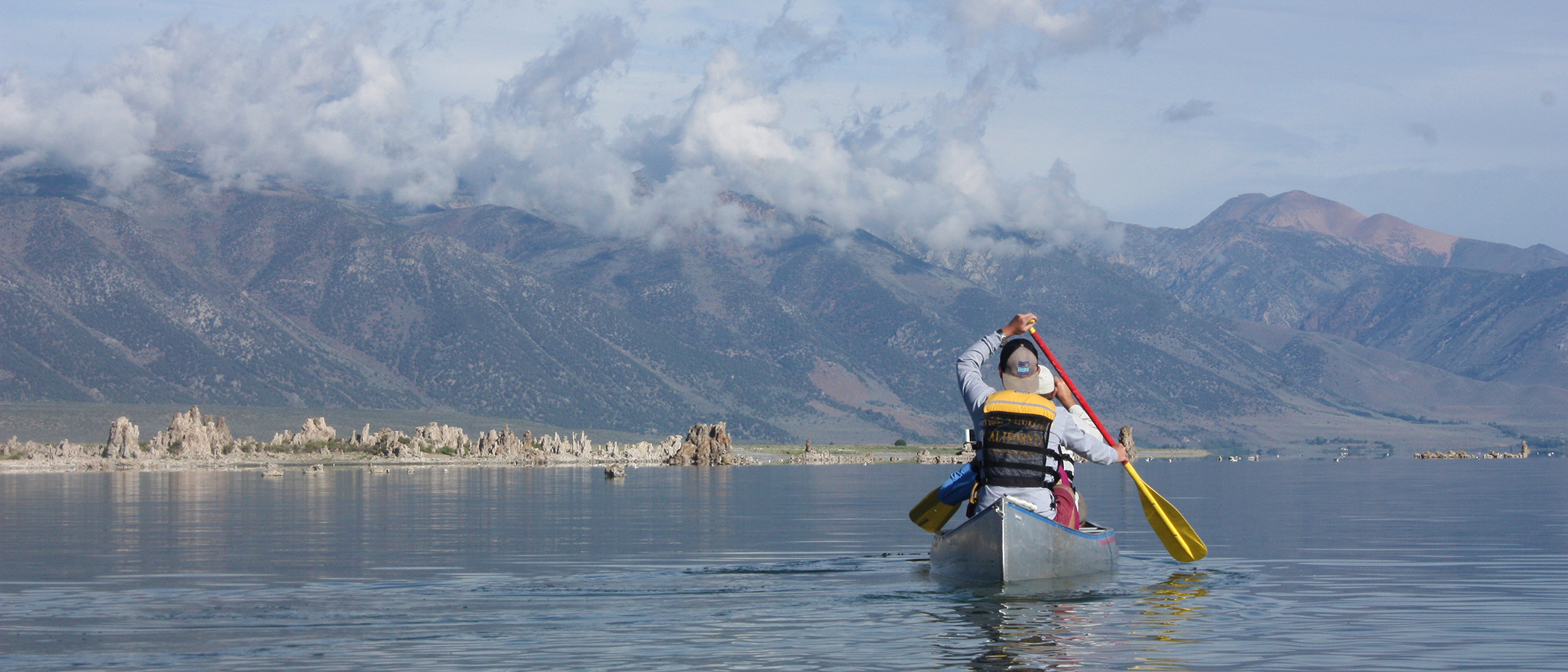 View from behind a person paddling a canoe on Mono Lake with tufa towers and tall mountains shrouded in clouds in the distance.
