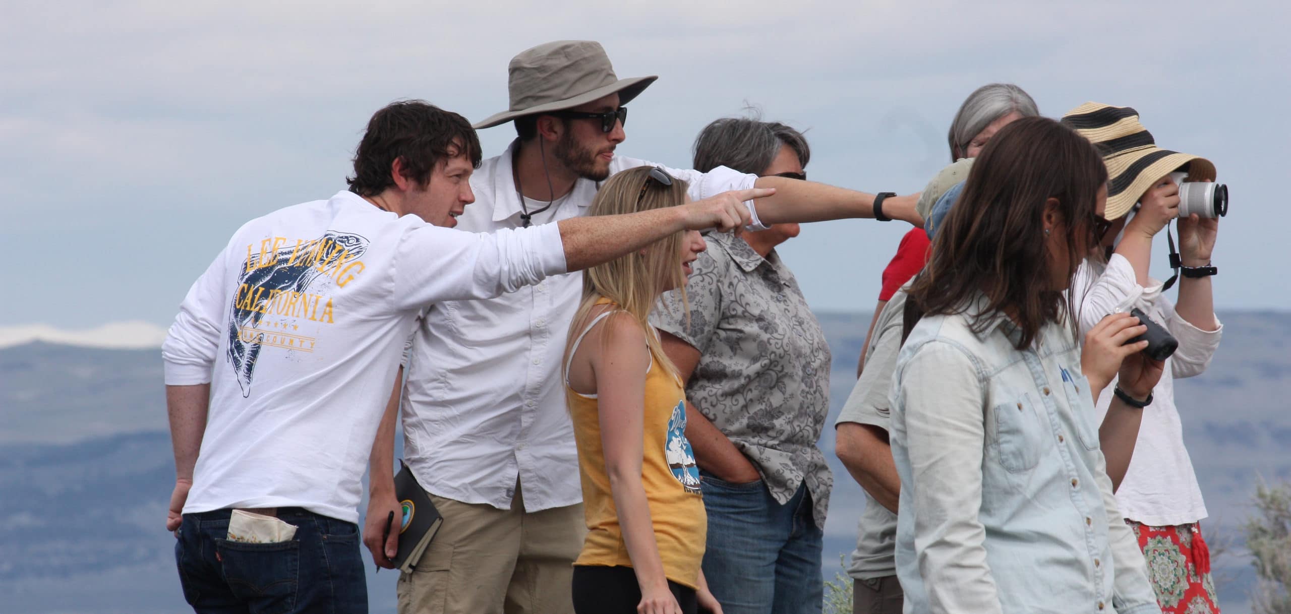 Nine people are looking at something interesting off in the distance, that we cannot see, and two people are pointing it out to one of the people, one is looking with binoculars, and one person is taking a photograph.