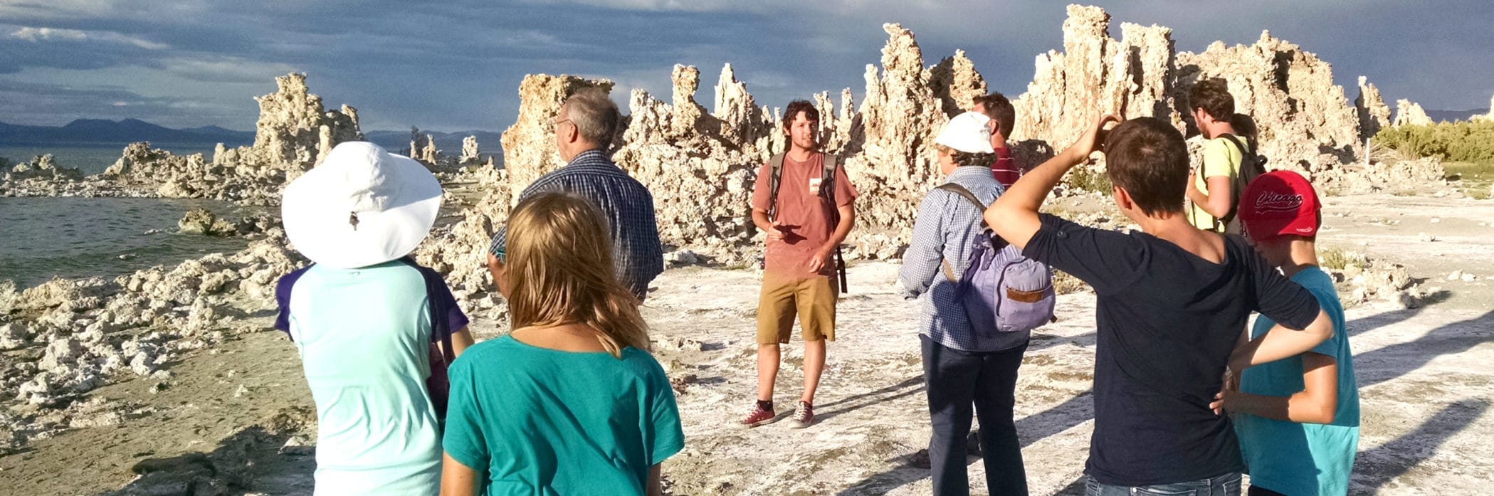 Tour guide talking to nine people at the shore of Mono Lake with tufa towers in the background, dramatic storm clouds, and the shoreline nearby.