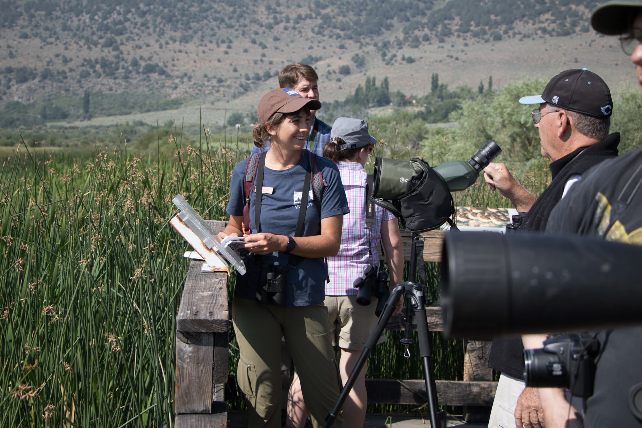 Five people with binoculars and spotting scopes are gathered on a wooden platform among marsh reeds happily doing some bird watching on a bright sunny day.