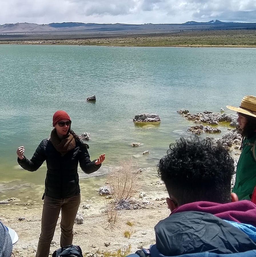Eleven people gather around a woman giving a tour at the shore of Mono Lake with the lake water in the background and the tour participants bundled up in warm outdoor gear.