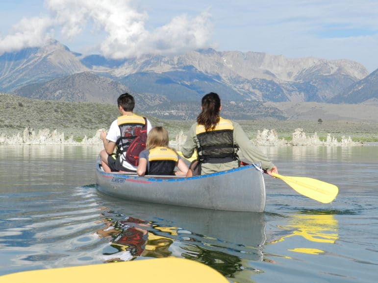 Three people sitting in a canoe paddling on Mono Lake in the bright sun moving over glassy water towards a grove of tufa towers with the tall Sierra Nevada in the distance.