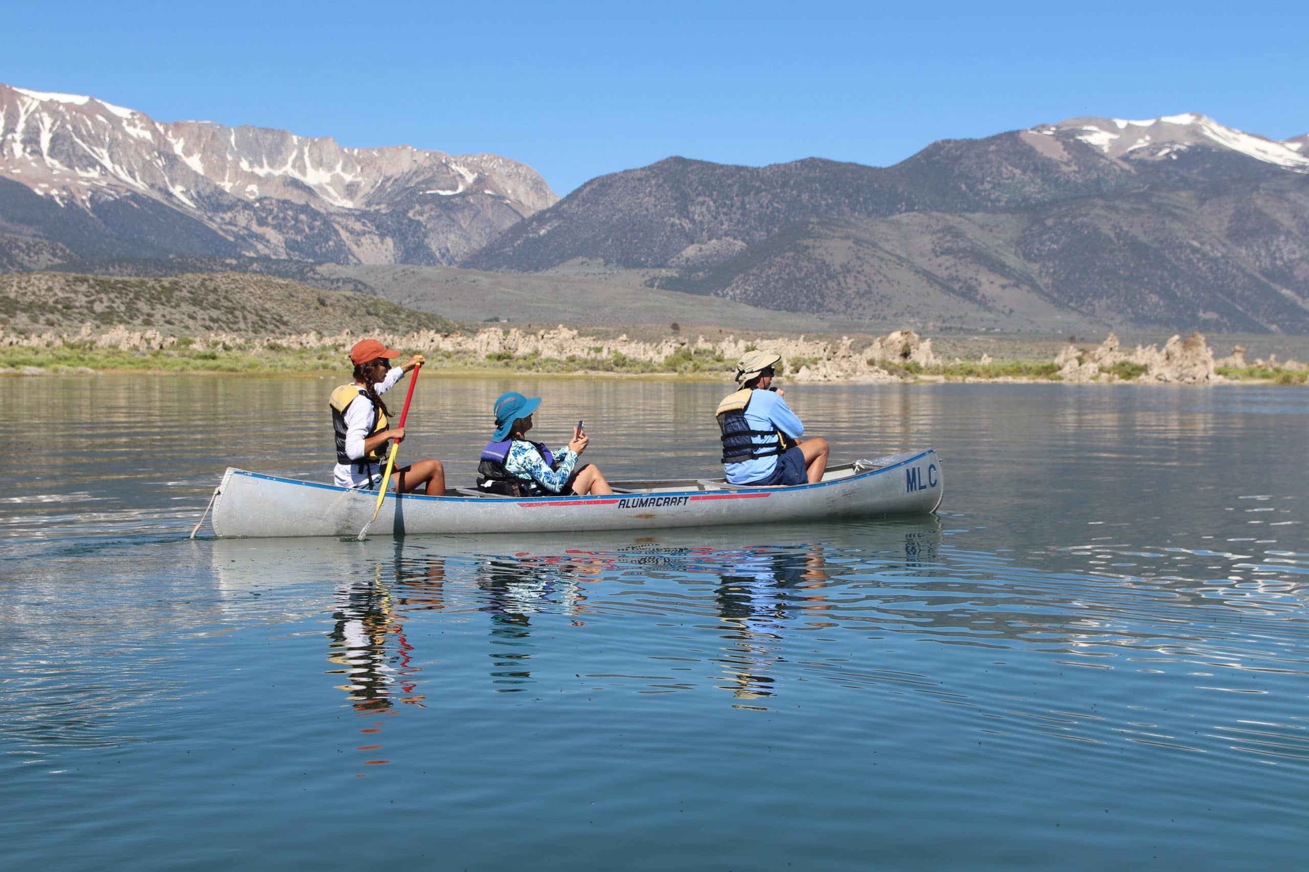 Three people canoeing on Mono Lake on glassy water with tufa towers and tall mountains with snow on them in the distance.