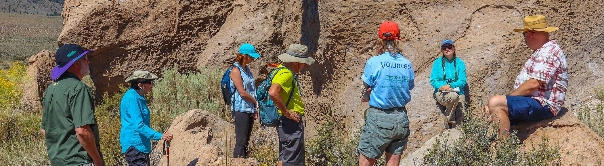 Seven people wearing hiking gear gather around a guide leaning on an unusual rock in a rocky and sunny outdoor setting.
