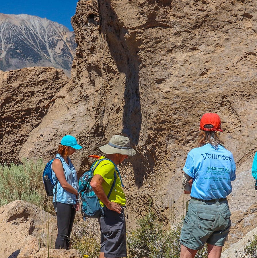 Seven people wearing hiking gear gather around a guide leaning on an unusual rock in a rocky and sunny outdoor setting.