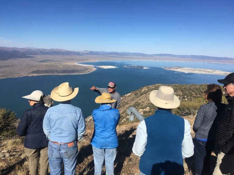 Six people gather around a guide who is using his hands to describe something, and the group is standing on a high precipice overlooking Mono Lake stretched out in a large expanse of blue with a clear view of the islands in the lake and the desert all around.