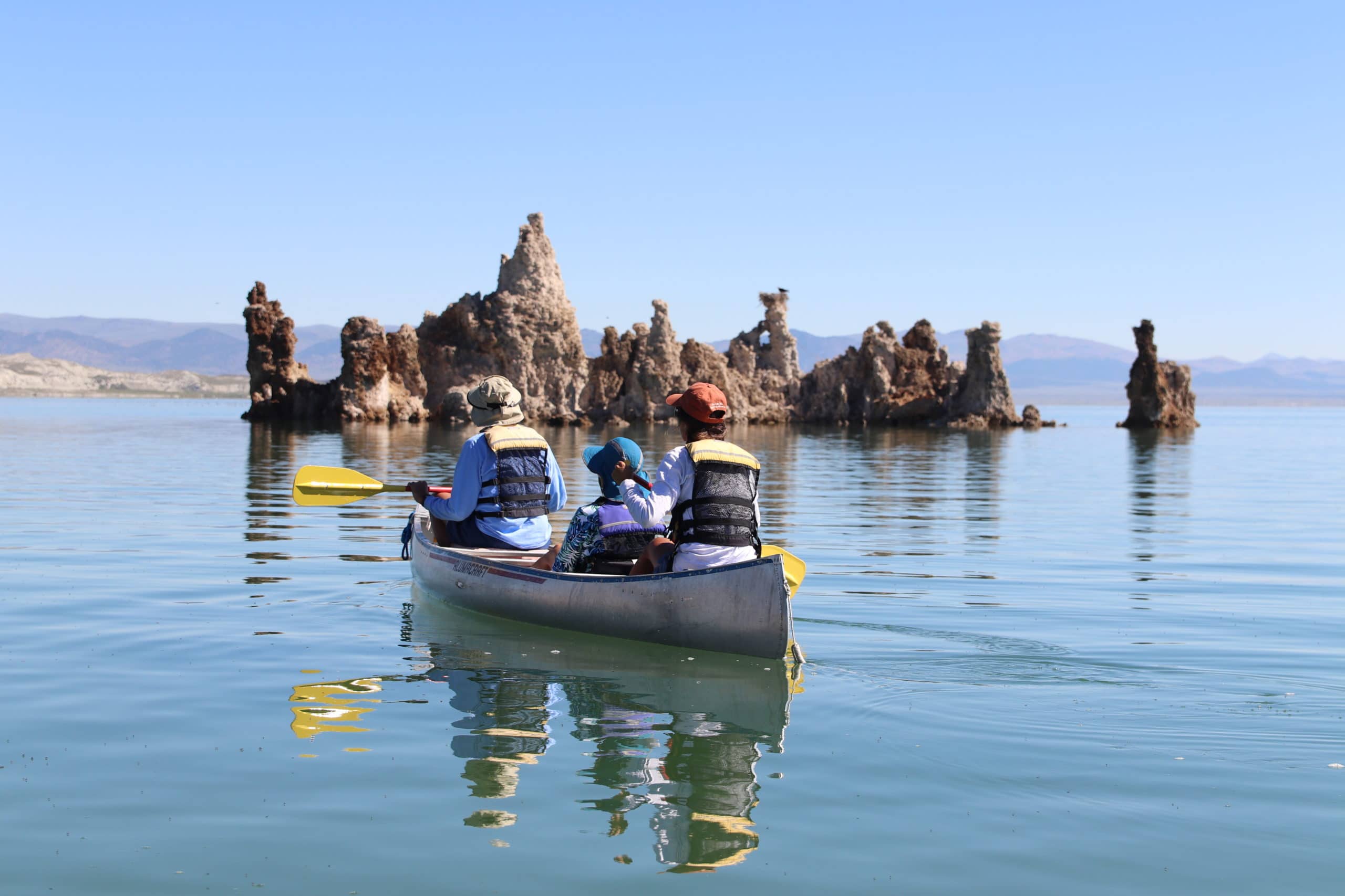 Three people canoeing on a bright sunny day on glassy Mono Lake towards an island of tufa towers with a large stick nest atop one of the towers and a fish-eating hawk, Osprey, perched on top.