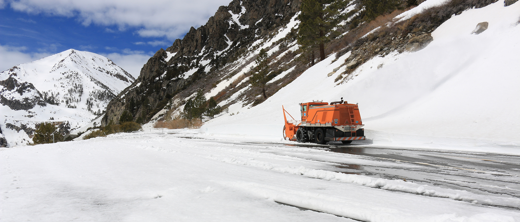 Orange Caltrans show removal vehicle next to a tall bank of snow with snow in the foreground.