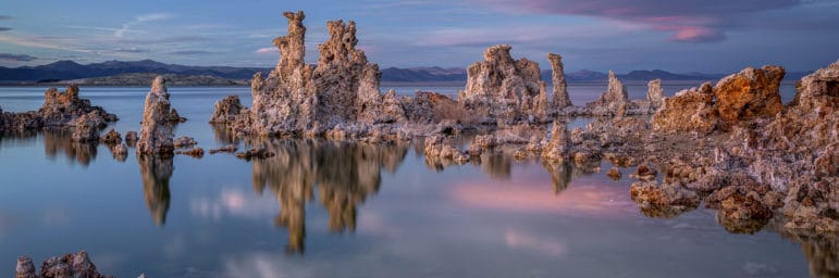 Tufa towers emerging from Mono Lake at sunset with blue skies and unusual clouds lit up in soft pink with beautiful soft light on the tufa towers reflected in the lake's surface, and you can even see some of the rocky shoreline under water.