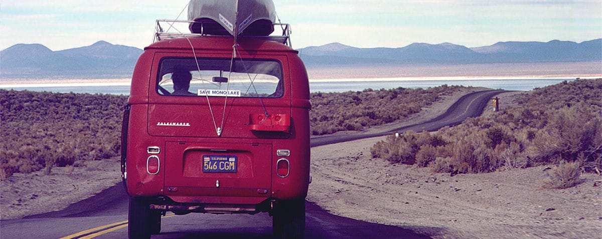 A bright red Volkswagen van with a metal canoe strapped on top and a "Save Mono Lake" bumper sticker drives down a two-lane highway towards Mono Lake in the distance.