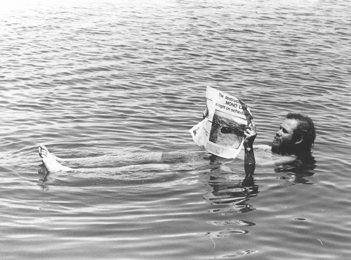 A bearded man, David Gaines, co-founder of the Mono Lake Committee floats on his back in Mono Lake while holding a newspaper that reads" The destruction of Mono Lake is right on schedule"--a fun publicity stunt.