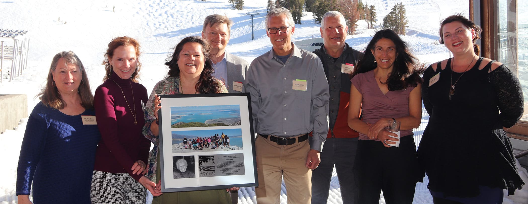 A group of 8 smiling people pose, one is holding a framed award with photos on it.