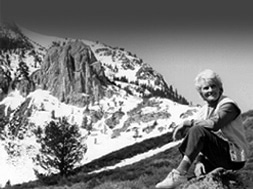 Black and white image of Andrea Mead Lawrence sitting on a rock with the craggy rocks and snow of the Sierra Nevada behind her.