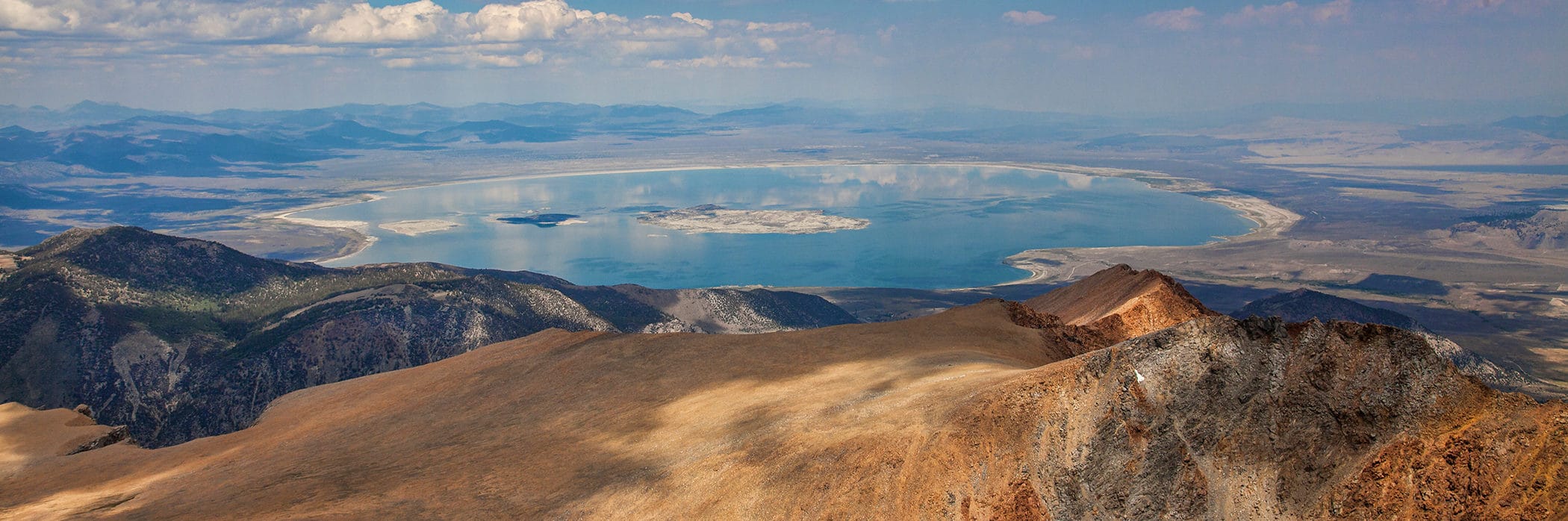 View of almost the entire Mono Lake from a tall mountain peak looking east out over the Mono Basin with Mono Lake's blue glassy reflection of clouds in the sky.