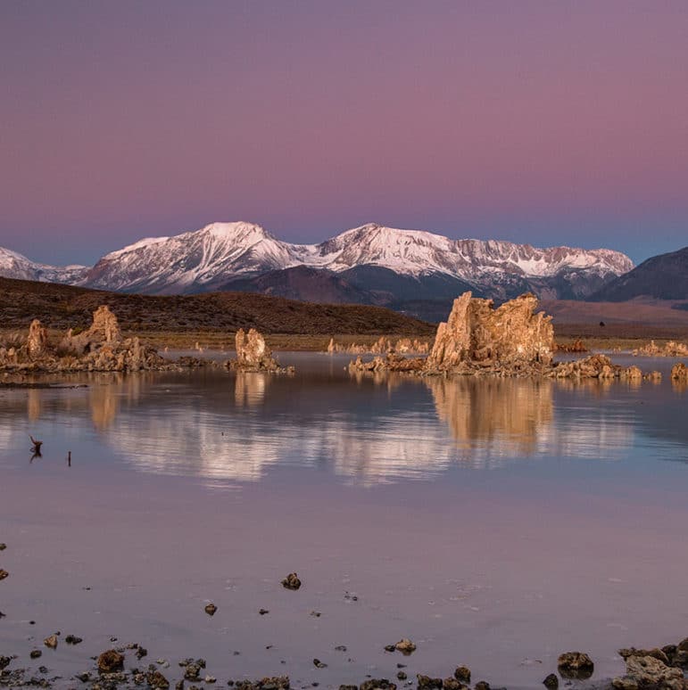 Shoreline of Mono Lake looking west towards the Sierra Nevada with tufa towers reflecting on the glassy lake and the sky is pink, purple, and blue.