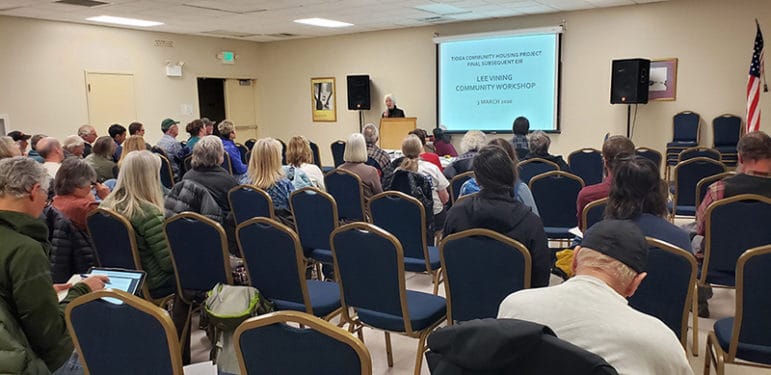 Fifty or so people sit in a community center looking up at a person giving a presentation into a microphone at a lecturn in front of a screen that reads "Tioga Community Housing Project Final Subsequent EIR, Lee Vining Community Worksshop".