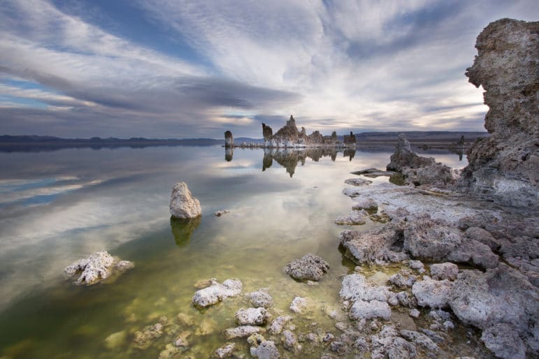 Dramatic scenic view of the shoreline of Mono Lake with a tufa tower and tufa rocks submerged in a green lake and large high clouds racing across the sky above an island of tufa towers perfectly reflected in Mono Lake.