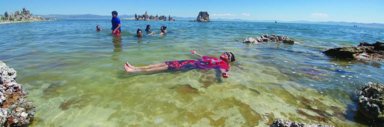 Six kids swimming in Mono Lake, with one kid floating on their back with goggles on, while surrounded by tufa rocks and with tufa towers in the distance.