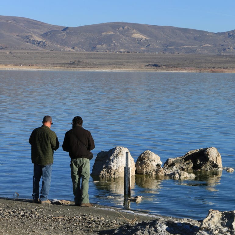 Two people stand next to each other on the shore of Mono Lake and seem to be conferring as they stand in front of a lake level gauge.