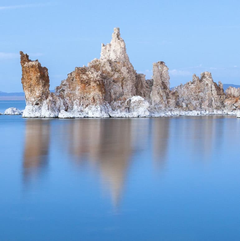 An island of white and rusty-brown tufa stands above the brilliant blue silky water of Mono Lake with a subtle blue sky.