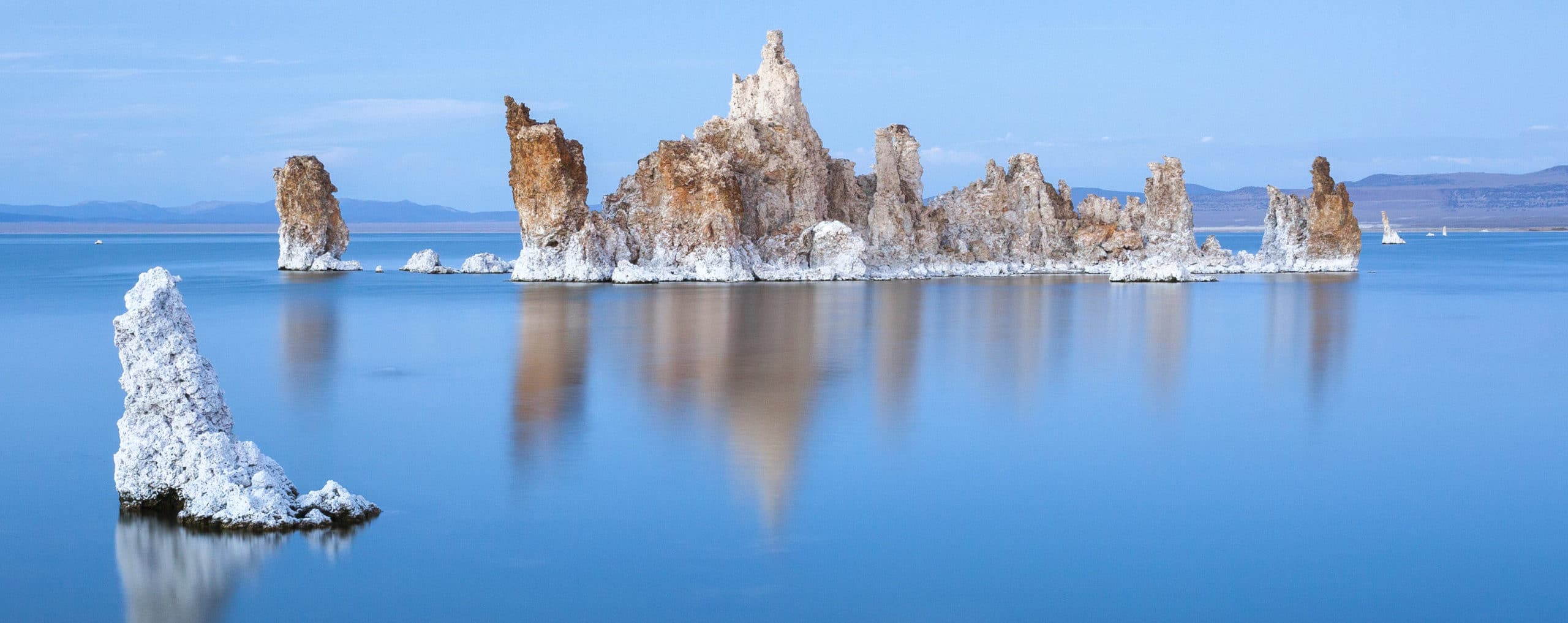 An island of white and rusty-brown tufa stands above the brilliant blue silky water of Mono Lake with a subtle blue sky.