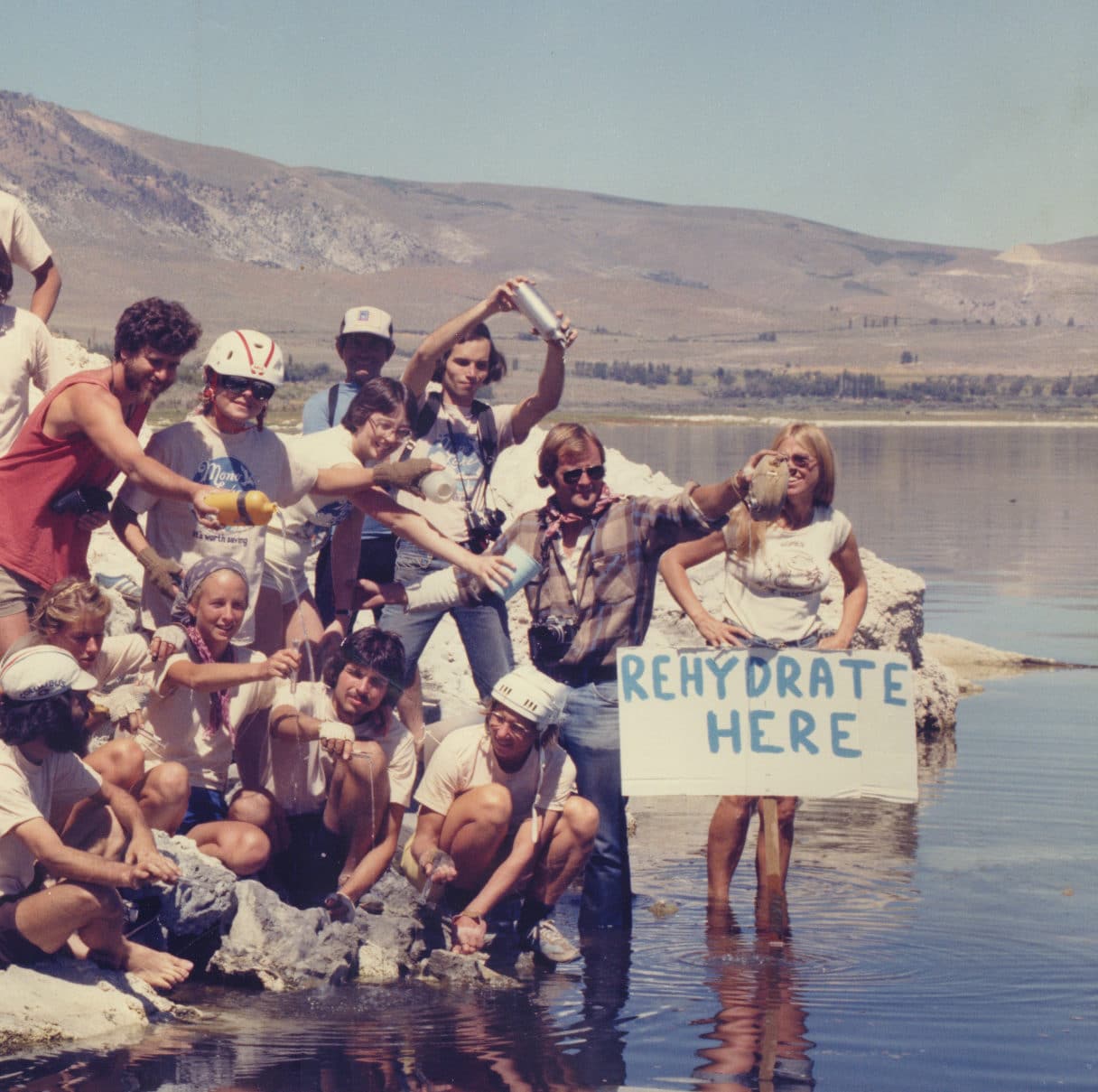 Group of about 50 people with hand-painted signs and Mono Lake T-shirts and many with bike helmets on pouring water from bottles and vials into Mono Lake.