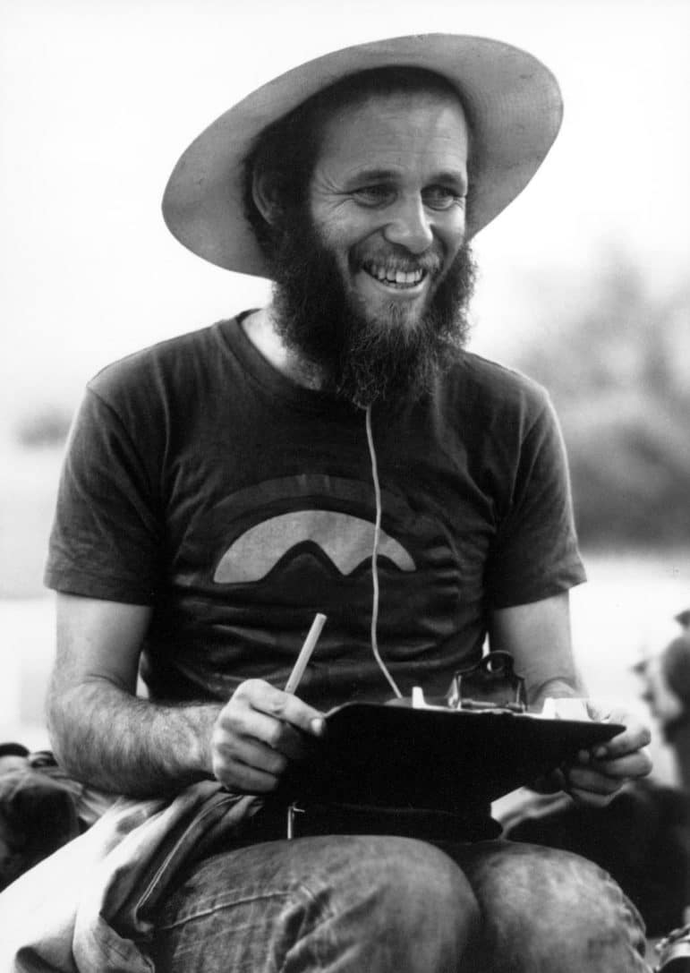 Black and white image of David Gaines with a beard, a hat, and a clipboard, with a big smile on his face.