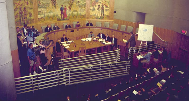 A hearing room with people seated in a semicircular room with reporters with large video cameras and a witness at the witness stand in front of a map display with Mono Lake on it.