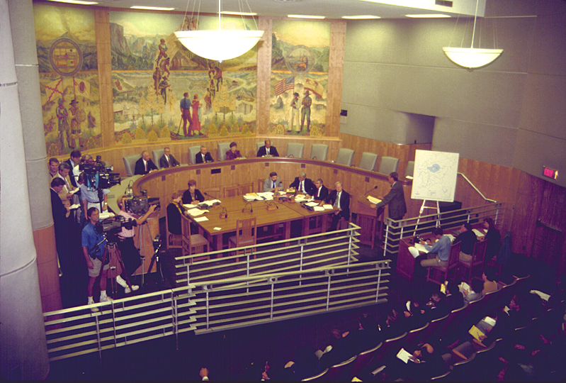 A hearing room with people seated in a semicircular room with reporters with large video cameras and a witness at the witness stand in front of a map display with Mono Lake on it.