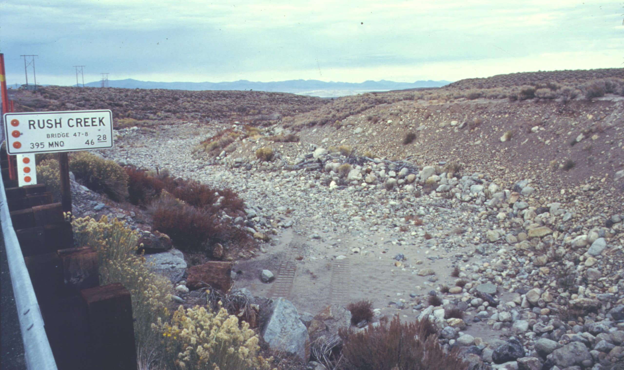 A dry creek bed with a highway sign that reads "Rush Creek" and motorized vehicle tracks in the sand of the creek below.