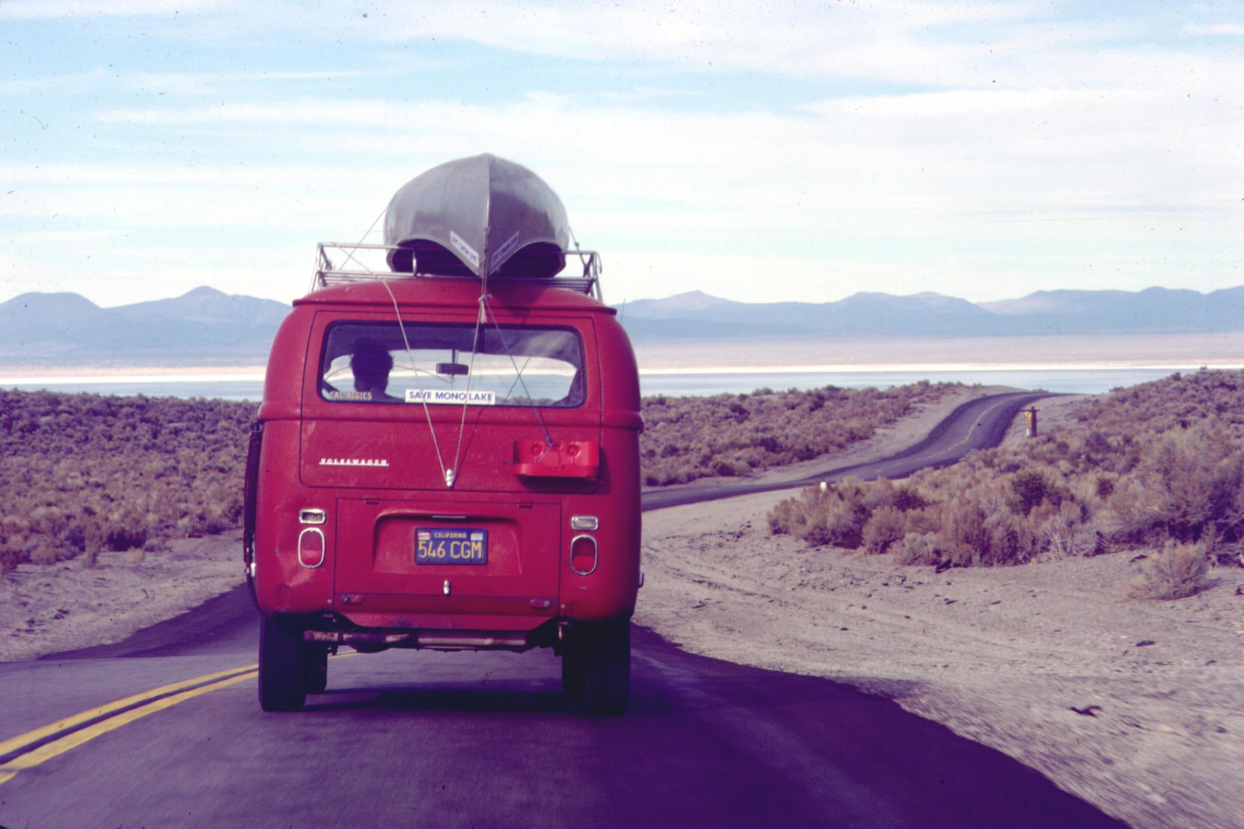 A bright red Volkswagen van with a metal canoe strapped on top and a "Save Mono Lake" bumper sticker drives down a two-lane highway towards Mono Lake in the distance.