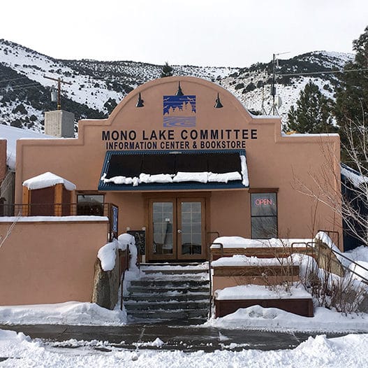 The storefront facade of the Mono Lake Committee Information Center & Bookstore after a recent snow, with snow piled on benches and solar panels but shoveled from the sidewalk and steps up the the building with an open sign in the window.