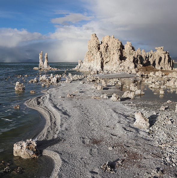 A storm brews in the distance behind a large grove of tufa towers on the shore of Mono Lake with a sandy peninsula dotted with smaller tufa mounds, in dramatic light, in the foreground.