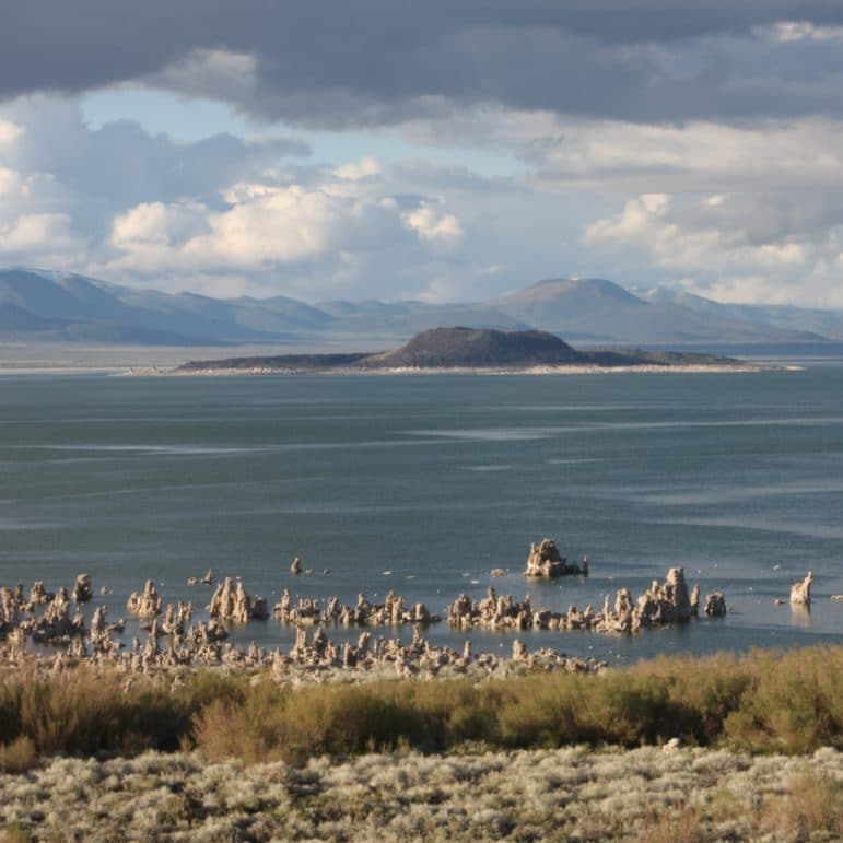 Black volcanic Negit Island in Mono Lake with tufa towers in Mono Lake in the foreground and hills in the background in muted hues of blue and grey with puffy clouds in the sky.