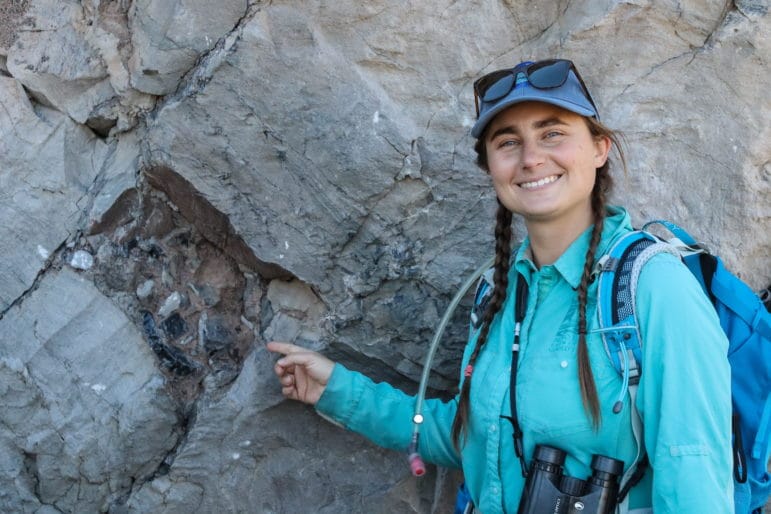 Woman with binoculars and outdoor guiding gear pointing out a geologic feature on a rock.