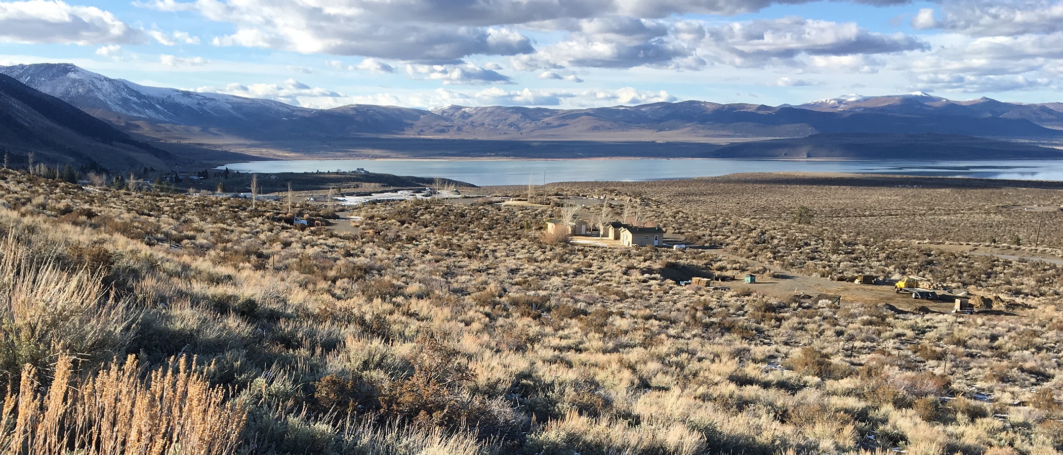 A few small houses sit in a dry green and yellow sagebrush field with a blue lake, dark mountains and a cloudy sky in the background.