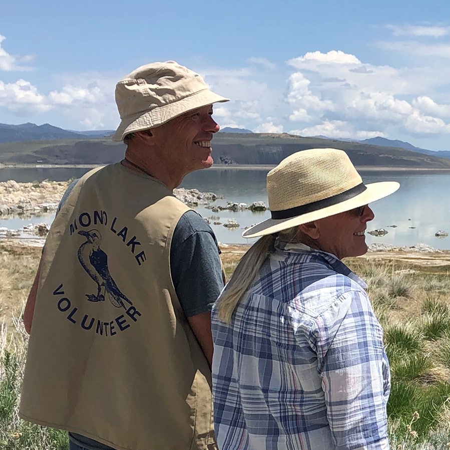 Four people overlooking Mono Lake on a bright sunny day smile at eachother while one looks through binoculars and tow of them have official Mono Lake Volunteer shirts and vests.