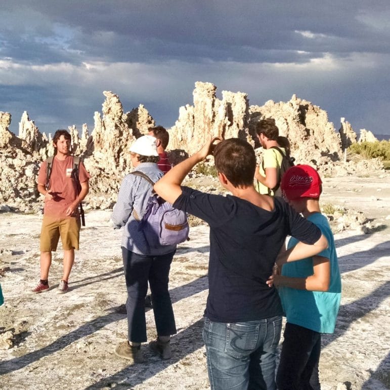 Tour guide talking to nine people at the shore of Mono Lake with tufa towers in the background, dramatic storm clouds, and the shoreline nearby.