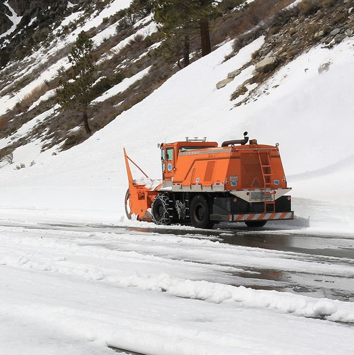 Orange Caltrans show removal vehicle next to a tall bank of snow with snow in the foreground.