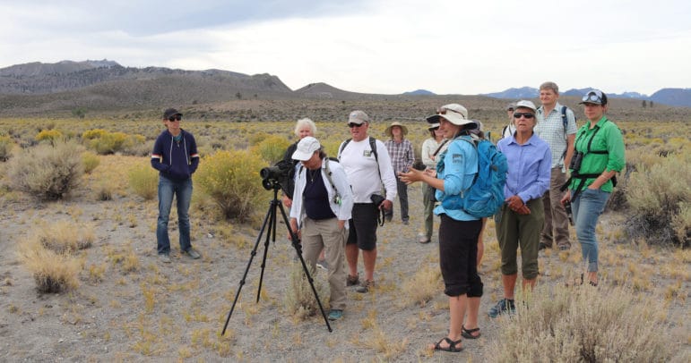 Twelve people gathered around a spotting scope and looking off into the distance on a sandy path in front of geologic craters in the background.