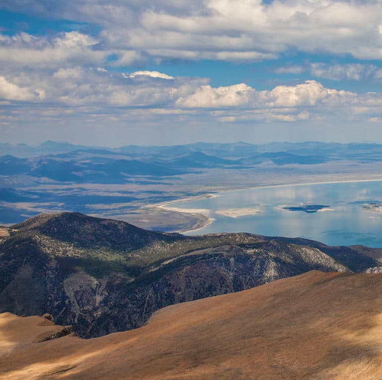 View of almost the entire Mono Lake from a tall mountain peak looking east out over the Mono Basin with Mono Lake's blue glassy reflection of clouds in the sky.