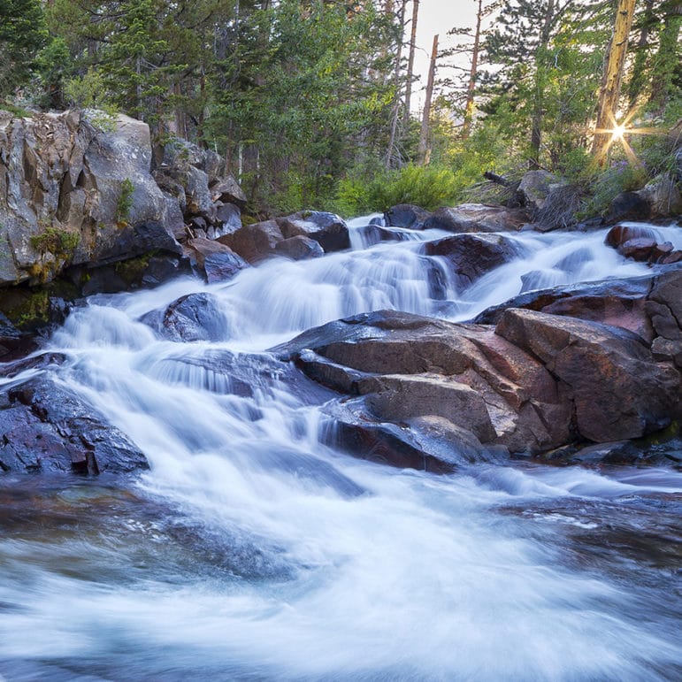 A long-exposure photograph of water running over rocks in a mountain stream so that the water looks white and silky as it passes over red and brown boulders before splashing out with motion into the foreground and you can see tall evergreen trees with bright green foliage above and sunlight making a golden-yellow sunburst through the forest.