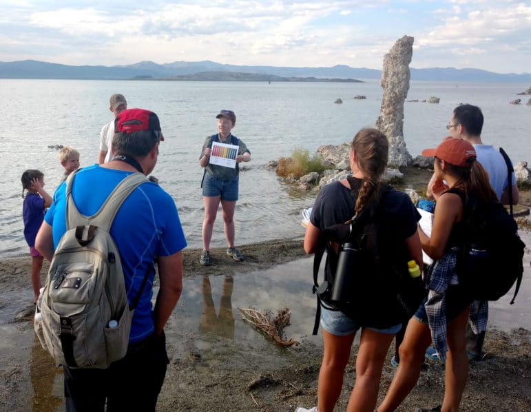 Eight people, including kids, gather around a tour guide holding a color chart while standing on a sandy strip of Mono Lake shoreline with a tall tufa tower and Mono Lake behind her as the group looks on.