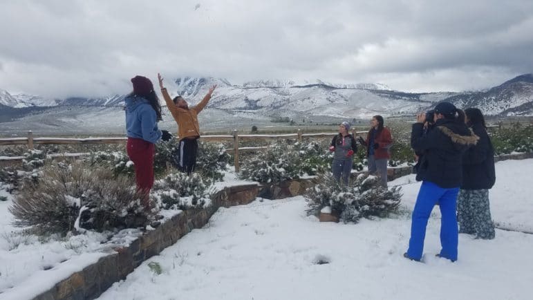 Six students stand in a circle outside in the snow with one jumping up and throwing a snowball in the air while the others laugh and take photos, with beautiful snowy mountains shrouded in clouds in the background.