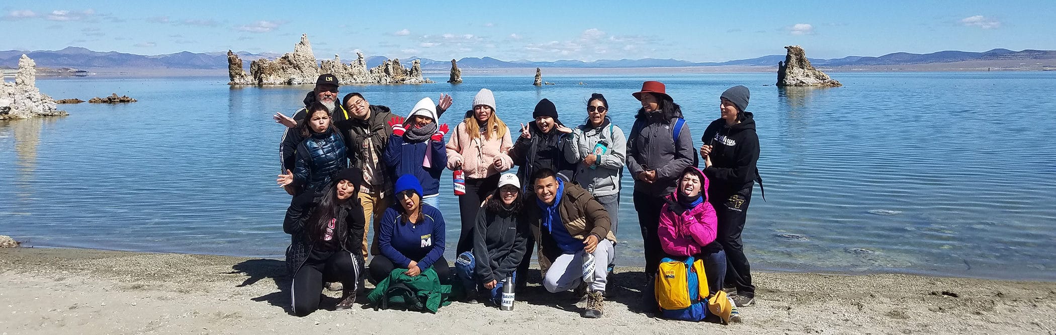 A group of students posing for a group photograph at the shoreline of Mono Lake with the blue lake in the background and tufa towers sticking up from the water and some of the students are making silly faces and gestures.