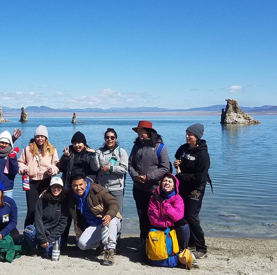 A group of students posing for a group photograph at the shoreline of Mono Lake with the blue lake in the background and tufa towers sticking up from the water and some of the students are making silly faces and gestures.