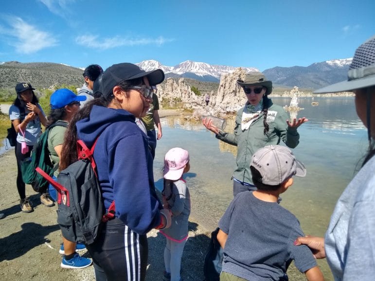 A tour guide gives a tour at the shore of Mono Lake with eight people, including kids, listening to her talk as she stands with tufa towers nearby and snowy mountains in the distance on a clear sunny day.