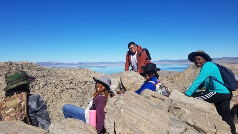 Five students on top of a rocky mountain in hiking gear looking out over Mono Lake under a clear blue sky, and they are looking happy and proud and relaxed.