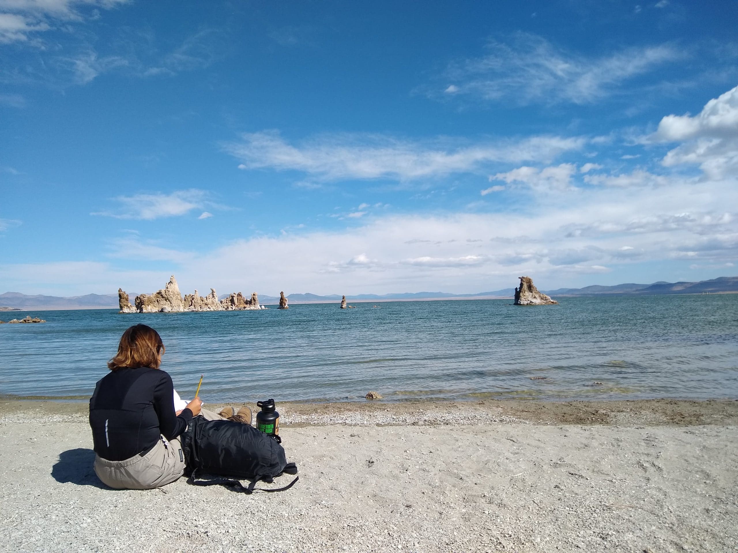 A student sits facing Mono Lake with her backpack as she writes in a journal with blue Mono Lake and an island of tufa towers and a blue sky with white clouds above.